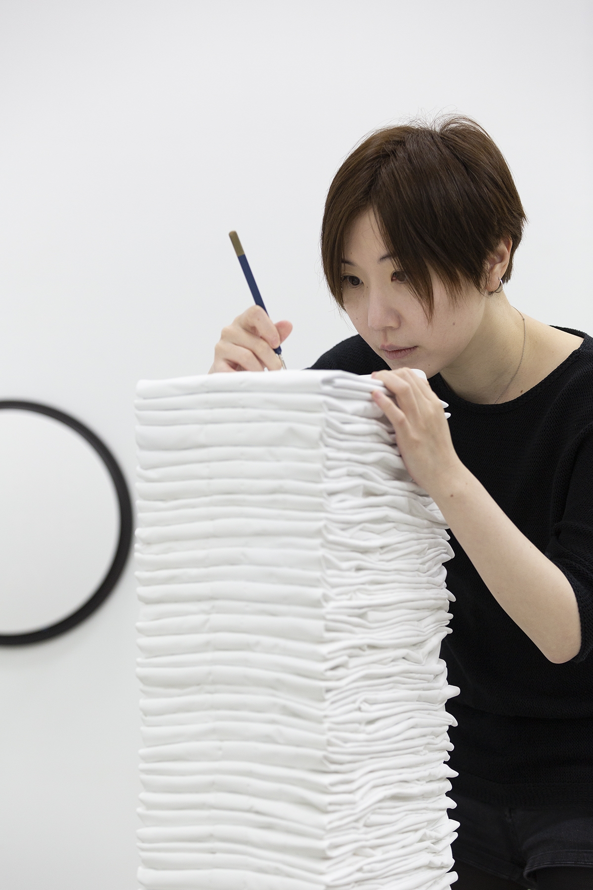 A person wearing a dark t-shirt leans on top of a stack of folded white fabric. She holds a pencil and uses it to mark the top of the fabric