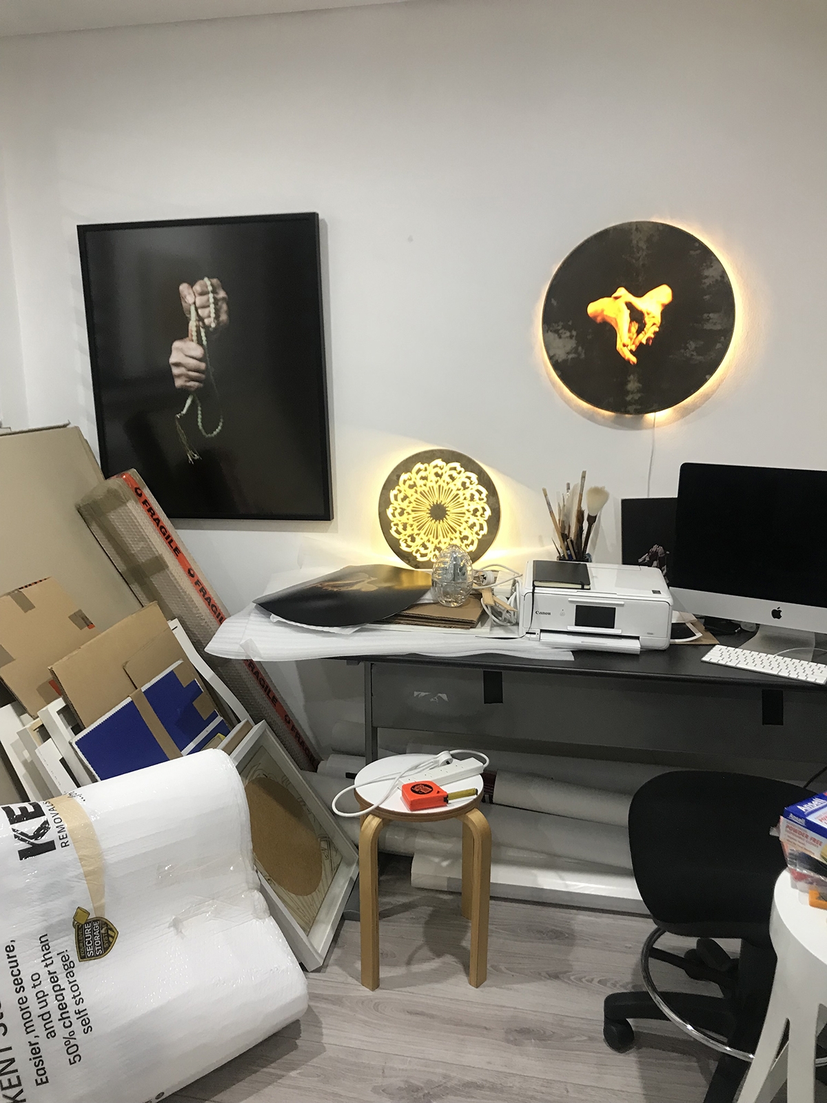 A framed photograph of hands, a stack of cardboard, an illumanited graphic artwork, a computer screen, and a stool are among the items seen in a white room.