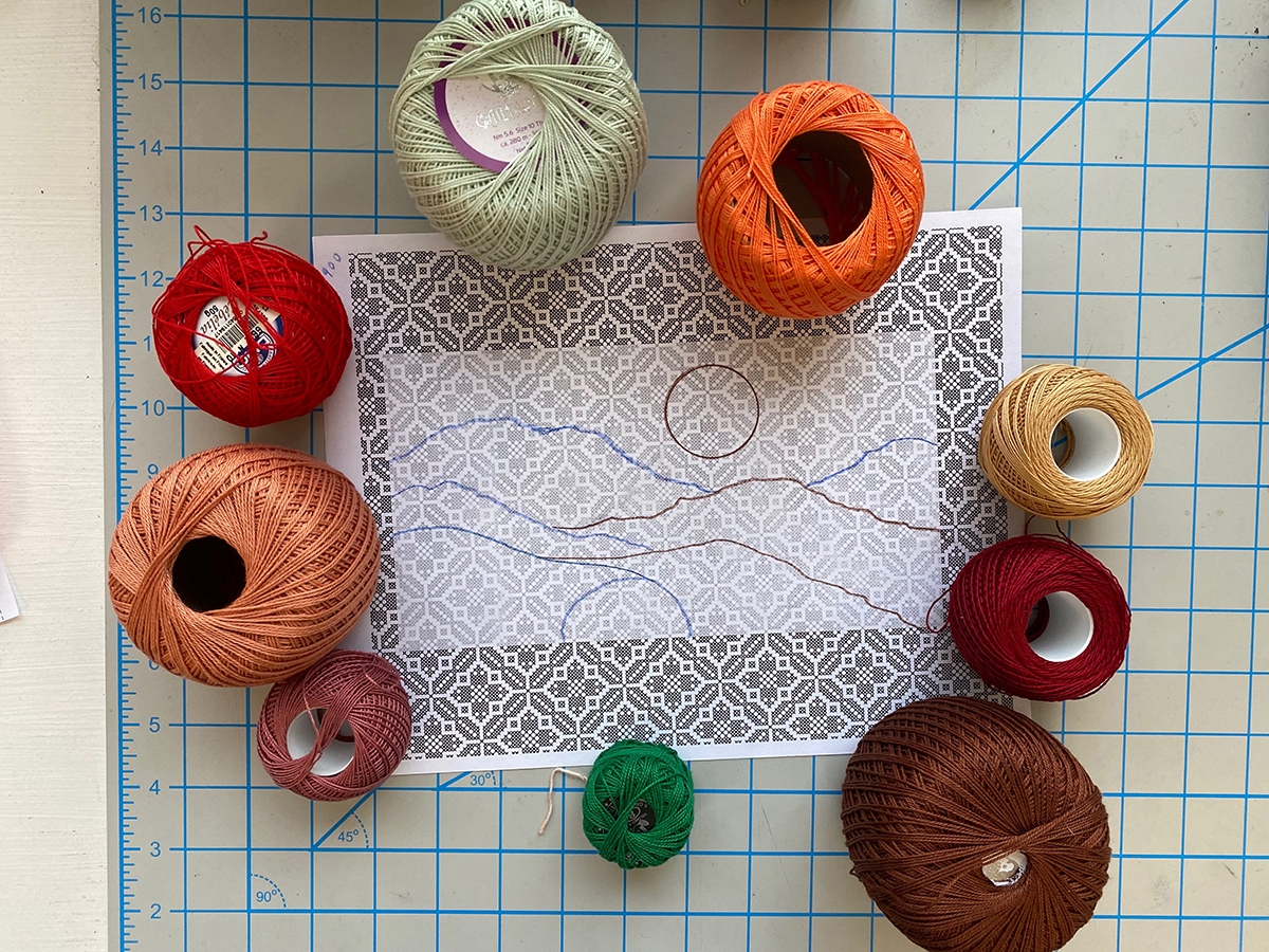Spools of colorful thread surround a line drawing of a landscape scene that sits on top of a drafting table