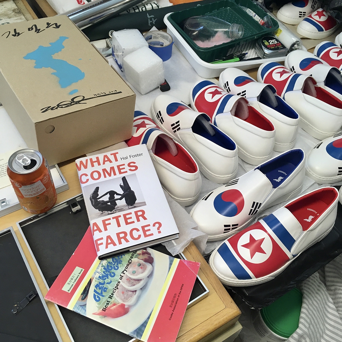 An orange aluminum can, a book by Hal Foster, a cookbook, and many pairs of white slip on shoes with the North and South Korean flags appear on a surface.