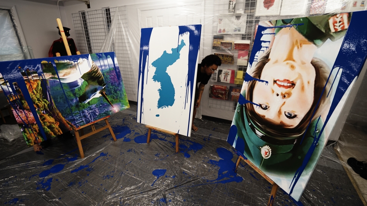 Large painted canvases inside an industrial studio. The canvas on the left depicts the Korean Peninsula. The canvas on the shows an upside down smiling face of a woman wearing a military-style cap.