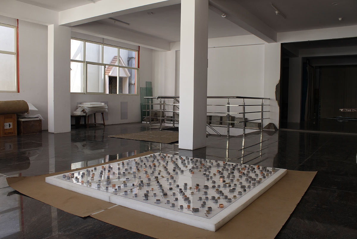 Hundreds of small block-shaped, metallic forms appear on top of a glass pane resting on a plinth on a shiny floor fo the artist's studio. 