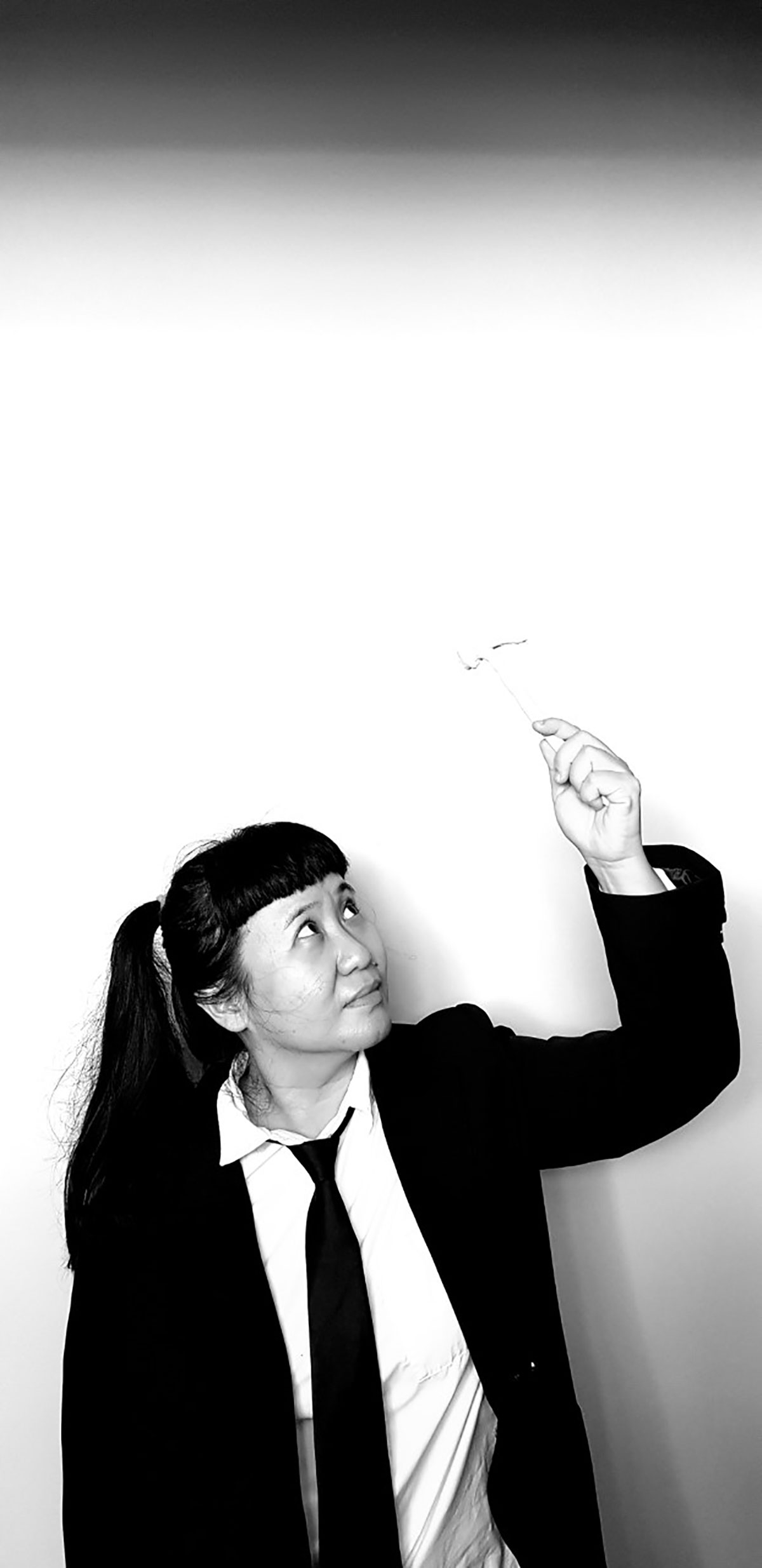A black-and-white portrait of anGie seah, who is dressed in a black blazer, white collared shirt, and black tie. Her long hair is in pigtails. She gazes upwards towards her raised hand, which is holding a miniature hammer.