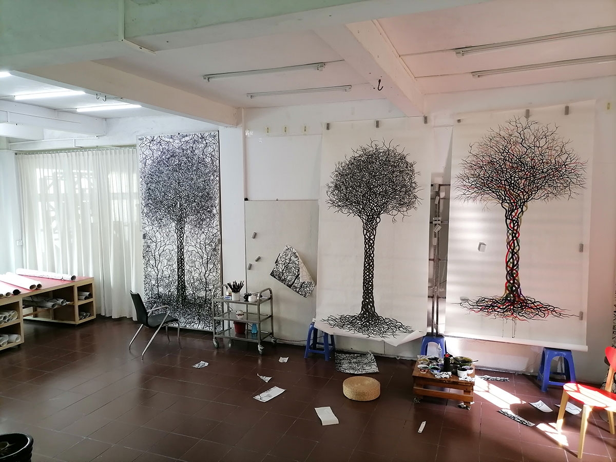 A photograph of the artist's studio. Three large, vertical sheets of paper are hung up; each features an intricately drawn tree with hundreds of interlaced branches and roots. A chair, drafting table, rolls of paper, paints, and paintbrushes are visible.