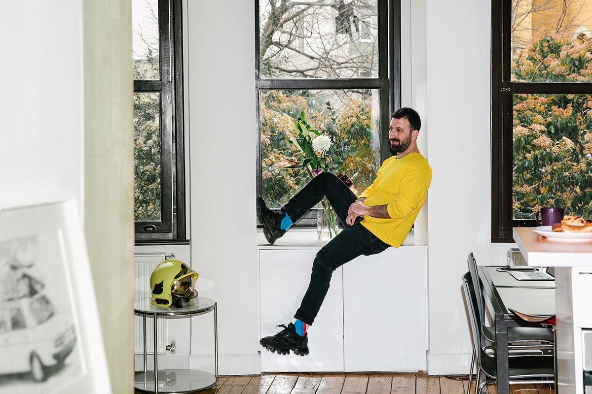 The artist sits in a windowill, smiling. He wears a yellow top and blacks pants and trees are visible outside the window.