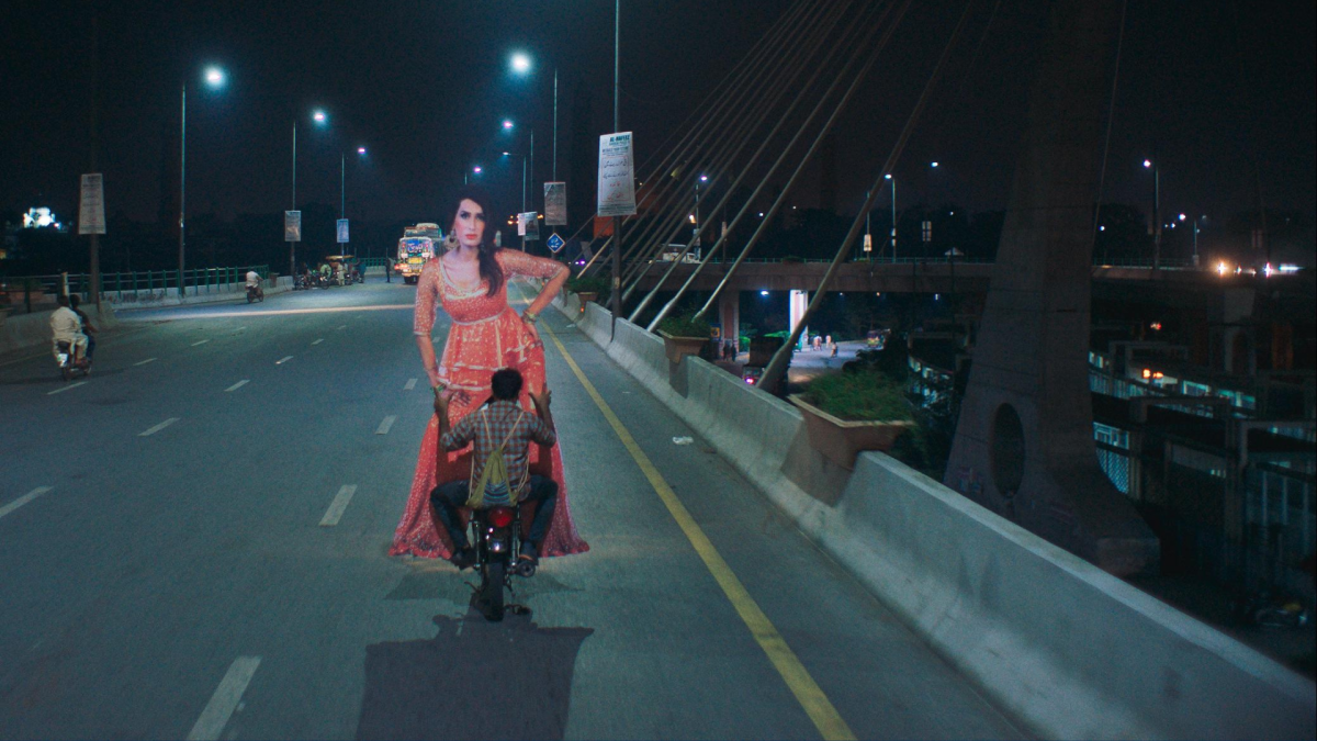 A man riding a motorcycle down a highway at night while carry a very large (twice his size) cardboard cutout of a dancer.