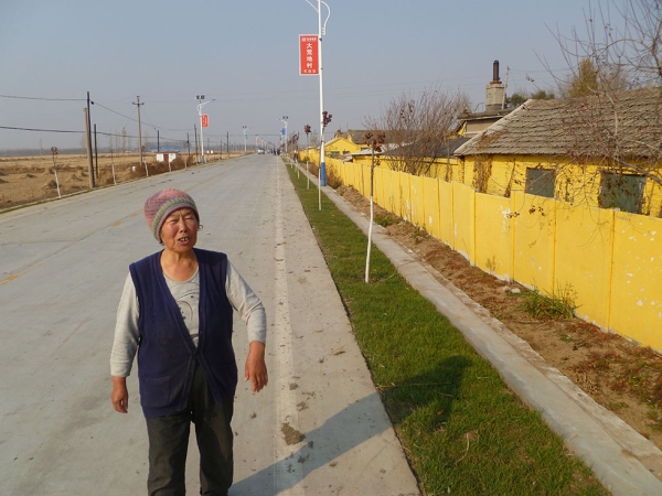 A local named Auntie Yi stands on the improved Red Flag Road, which now features solar-powered streetlights and Wasteland's first "lawn" — a strip of ornamental sod. (Michael Meyer)