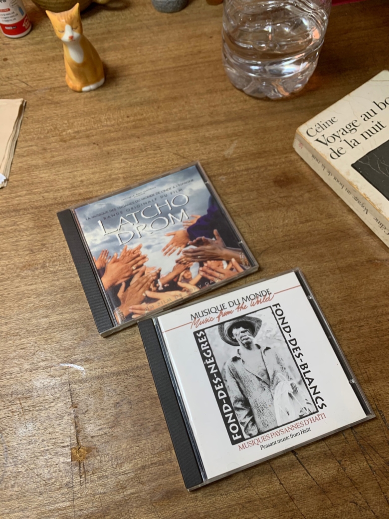 Two CDs appear on a table alongside a paperback book, a statuette of an orange cat, and a glass.