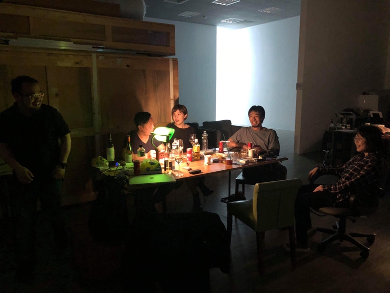 Five people are gathered around a table covered in bottles, glasses, and a computer.. They are smiling and laughing. 