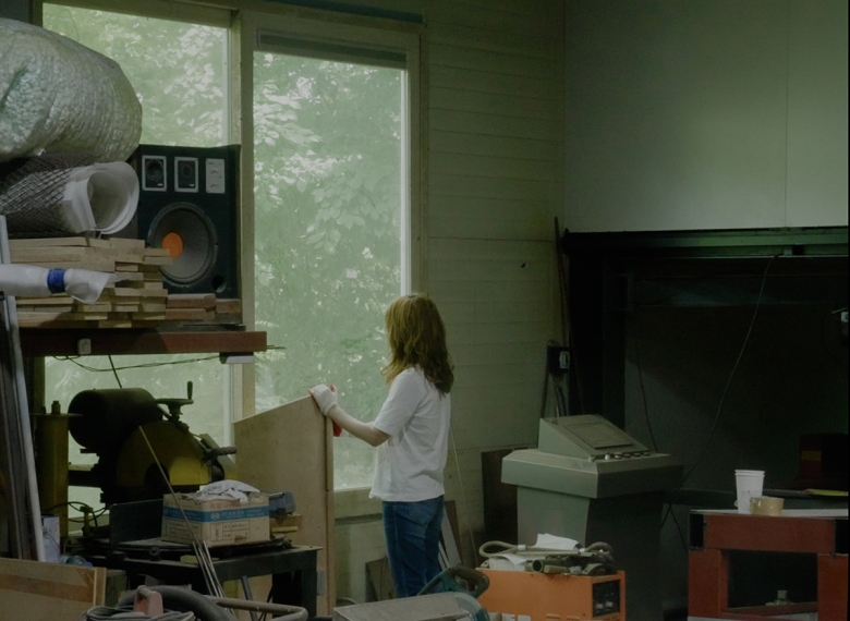 A person stands in an artists studio, looking out a window. She wears a white t-shirt, jeans, and white gloves. Lumber, cups, and boxes appear around her.