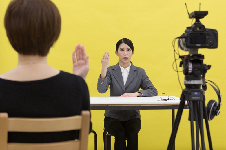 A person sits at a bare table, wearing a white-button down shirt and gray blazer. Her right hand is raised, the back of her hand facing the viewer. Another person sits across from her, also raising their right hand. They sit in front of a yellow background, and a video camera on a tripod appears at the right.