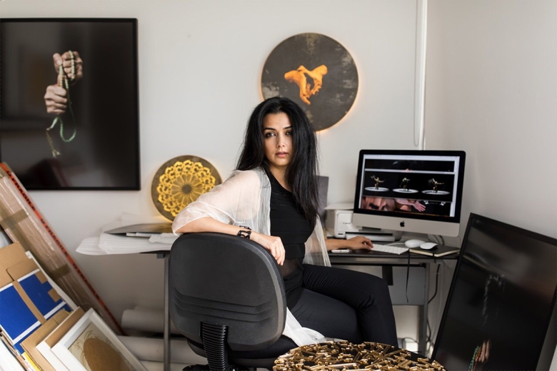Nasim Nasr sits in front of her computer. She wears a black shirt and pants with a sheer white jacket. She is surrounded by artworks featuring hands.