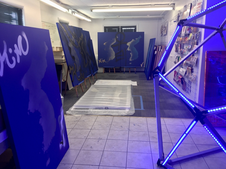 A series of large, blue canvasses are propped on easels around a tile-floored room. A bright, electric, blue sculpture appears at the right. 