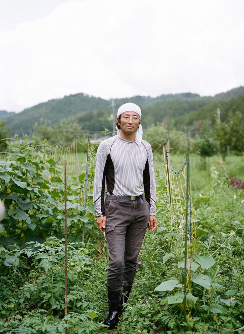 Ken Yonetani stands facing the camera. He is surrounded by lush green vegetation. He has a slight smile.