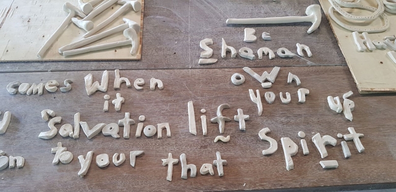 Letters made of clay appear on a wood tablet, spelling out words: Be, shaman, comes, when, own, it, salvation, lifts, your, up, to your, that, and spirit. Several hammers made of clay are on the table, as well.