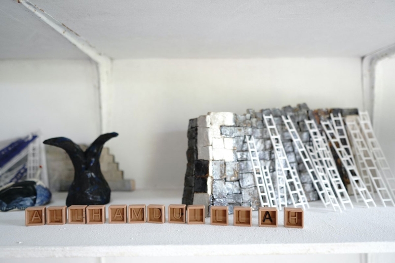 Small objects appear on a white shelf include several box-shaped pieces inscribed with a letter, miniature ladders, a black clay piece with a forked top, and black, white, and silver stacked blocks.