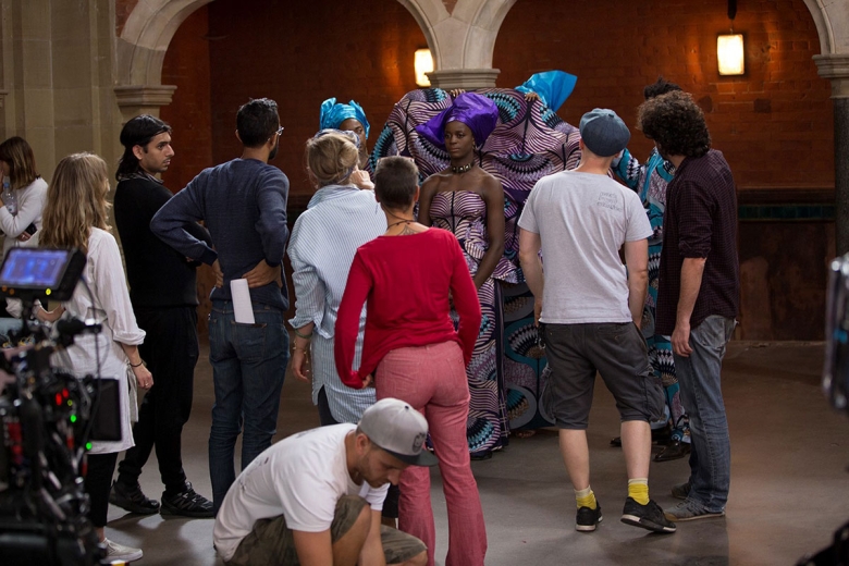Several people stand with their backs to the camera, facing a woman wearing an ornate dress and headwrap in the purple and teal patterns. Some film equipment is visible in the foreground. 