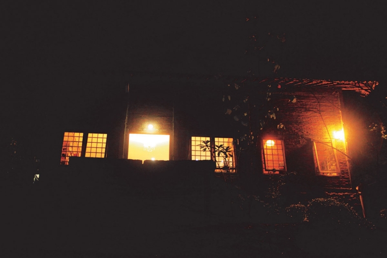 A building photographed at night. There are seven rectangular windows warmly lit. The silhouettes of trees and bushes are faintly visible.