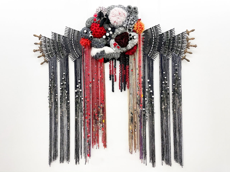 A hanging sculpture composed of yarn, rakes, woven belts, beads, and metal and plastic ornaments