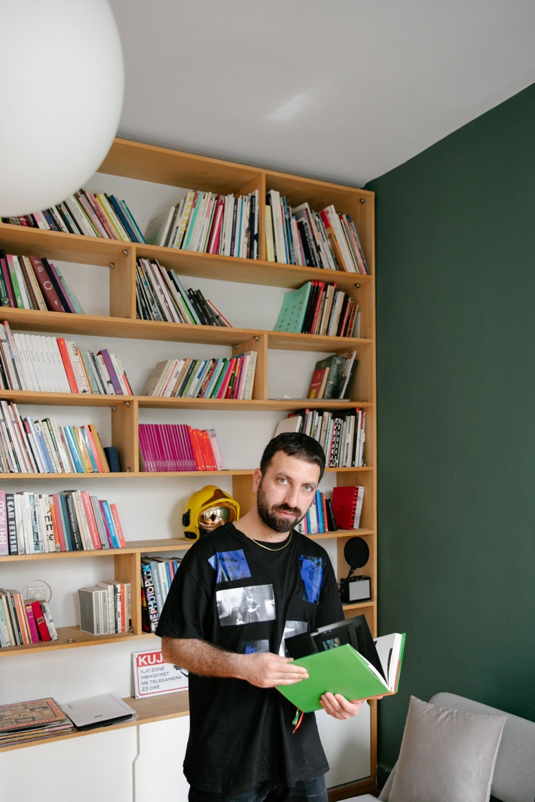 The artist stands in front of a bookcase, holding a green book and looking into the camera